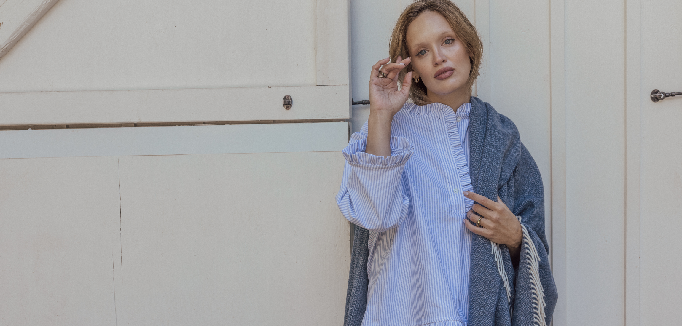 Woman wearing a light blue striped shirt and gray cardigan standing against a white wall.