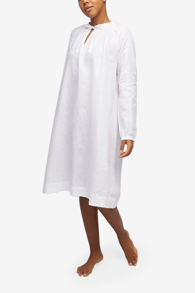 Full shot of a black woman with a blonde buzz cut,  wearing a knee-length nightgown with raglan sleeves with button cuffs. There is a little bow at the centre front of the gathered neck line. Made in our favourite, crisp white linen.
