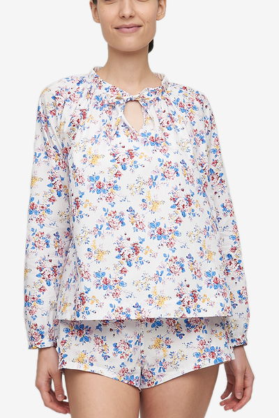 Cropped shot of a white woman, her torso is the focus.She is wearing a pyjama top with raglan sleeves with button cuffs, the hem hitting her at the hip. There is a little bow at the centre front of the gathered neck line. Made in a woven cotton with red, blue and yellow vintage florals.