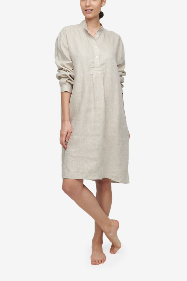 Full shot of a white woman with her hair tied back in a low bun, barefoot against a white background. She is wearing a below-the-knee-length nightshirt that has long, cuffed sleeves, a three-quarter placket with 4 buttons and a stand collar. It's made in cool beige, crisp, textured linen.