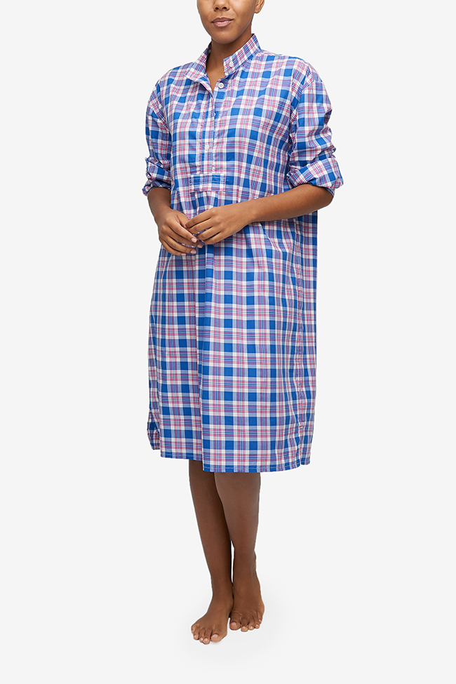 Full shot of a black woman with a blonde buzz cut, barefoot against a white background. She is wearing a below-the-knee-length nightshirt that has long, cuffed sleeves, a three-quarter placket with 4 buttons and a stand collar. It's made from a lightweight cotton shirting in a blue, red and white plaid.