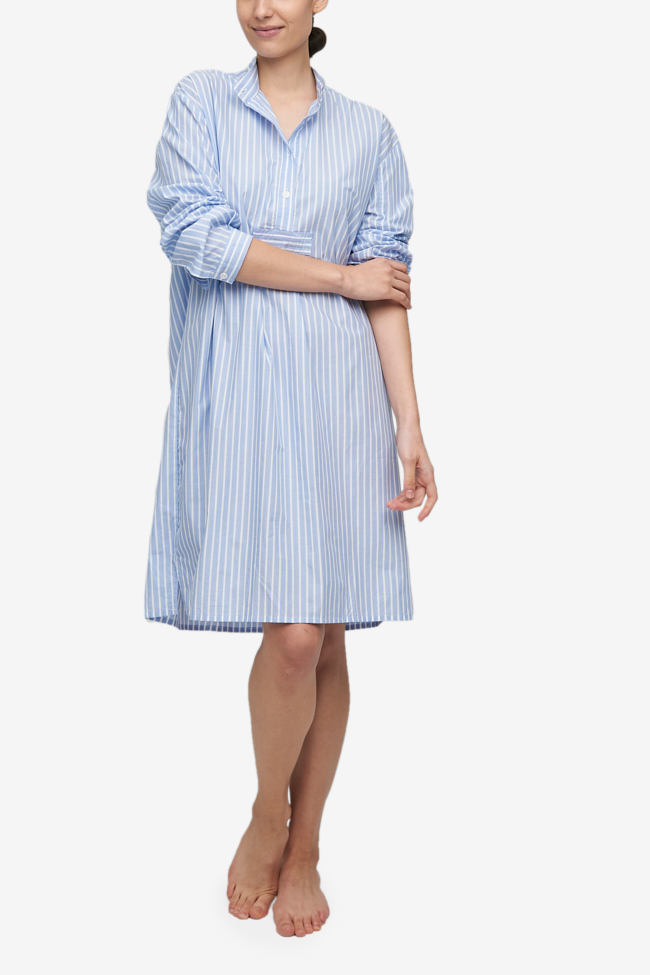 Full shot of white woman with her hair tied back in a low bun, barefoot against a white background. She is wearing a below-the-knee-length nightshirt that has long, cuffed sleeves, a three-quarter placket with 4 buttons and a stand collar. It's made from a lightweight cotton shirting with subtle blue and white vertical stripes.