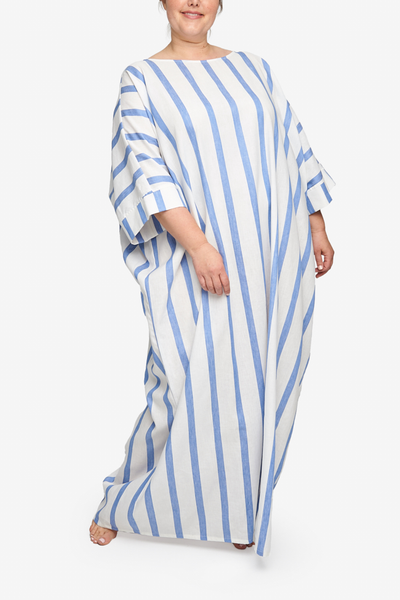 Full body shot of our plus-sized model wearing an oversized kaftan. It is floor length with a boat neck, and wide, three quarter length sleeves, of course pockets. It has an overly generous amount of ease, especially in this hips giving it a draped, extra relaxed fit.It's made from a soft and drapey white cotton linen blend with bold, blue vertical stripes.