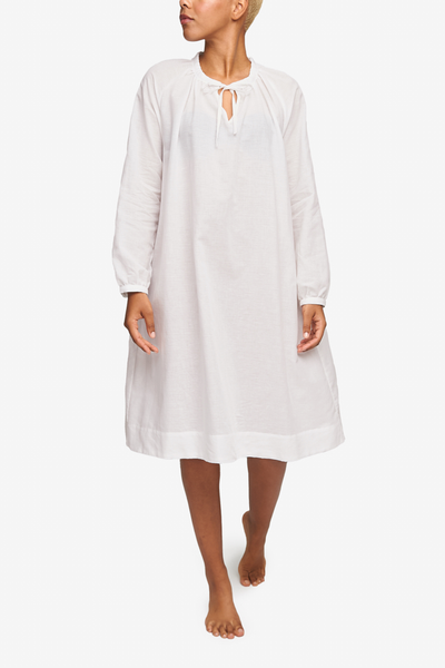 Full shot of a black woman with a blonde buzz cut. She's looking over her right shoulder as she walks toward the camera. Wearing a knee-length nightgown with raglan sleeves with button cuffs. There is a little bow at the centre front of the gathered neck line. Made in a ultra lightweight, sheer white cotton linen blend.