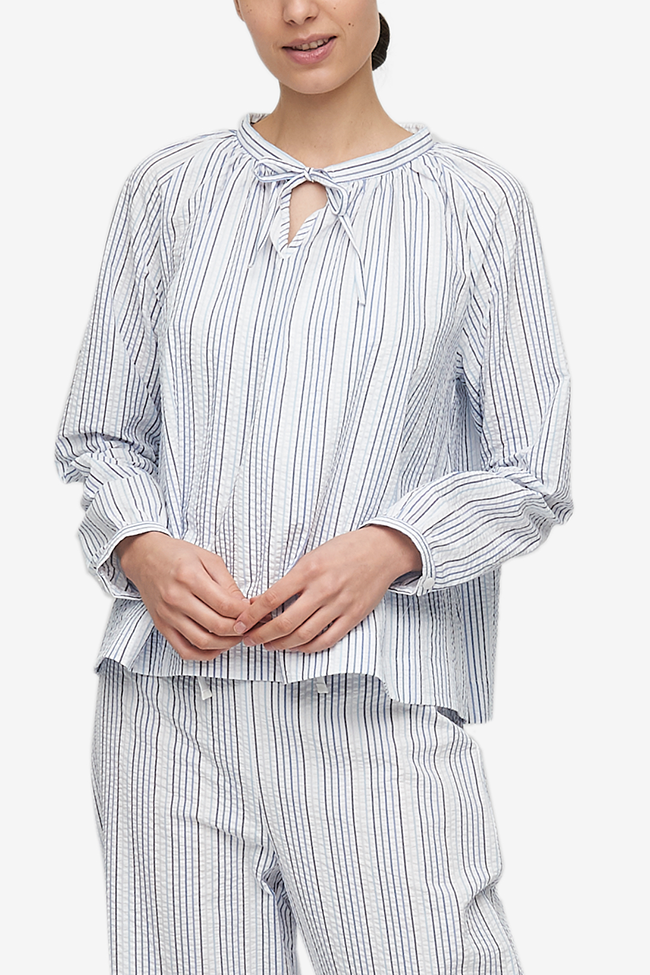 Cropped shot of a white woman, her torso is the focus.She is wearing a pyjama top with raglan sleeves with button cuffs, the hem hitting her at the hip. There is a little bow at the centre front of the gathered neck line. Made from a beautiful white seersucker cotton with thin blue stripes.