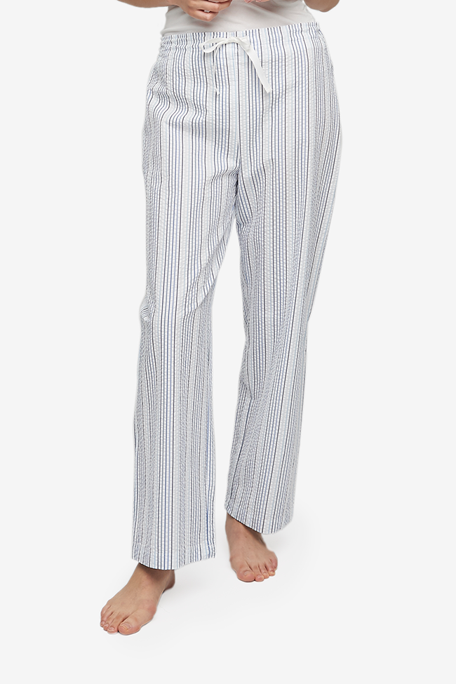 Cropped shot of a white woman, her lower body and legs are the focus. Wearing a wide leg pyjama pant with a twill tape drawstring front and elastic back waist band. It's made from a crisp, lightweight seersucker cotton with vertical blue stripes on a white base.