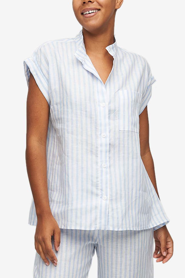 Cropped shot of a black woman, her torso is the focus. Wearing a pyjama top with a full placket and cuffed, cap sleeves. One breast pocket on the left side. It's made from a crisp linen with 3/4" blue and white stripes.
