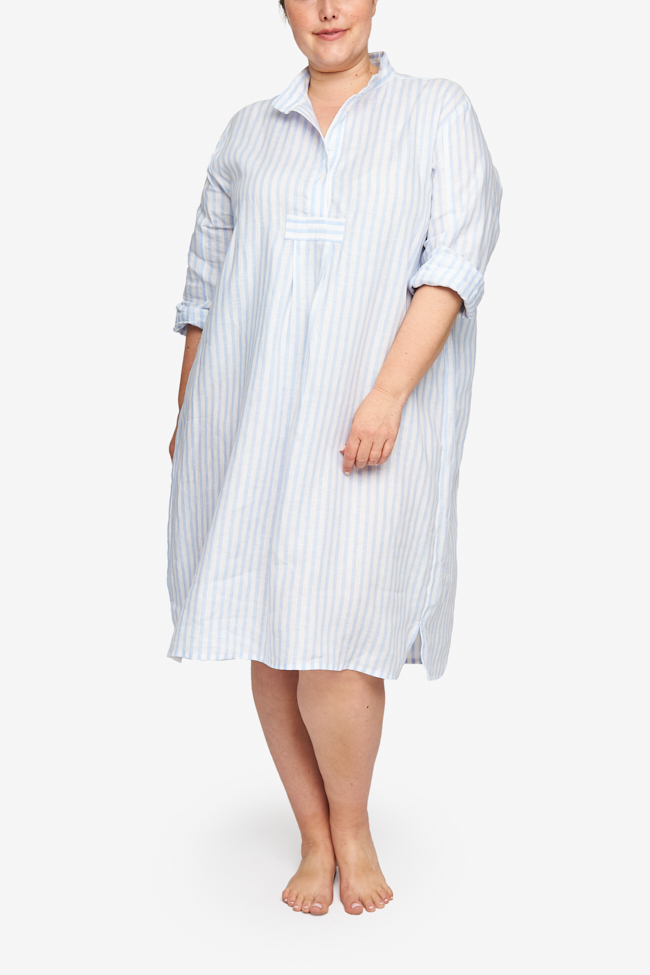 Full shot of a plus-sized white woman with her hair tied back in a low bun, barefoot against a white background. She is wearing a below-the-knee-length nightshirt that has long, cuffed sleeves, a three-quarter placket with 4 buttons and a stand collar. It's made in a white and blue striped linen.