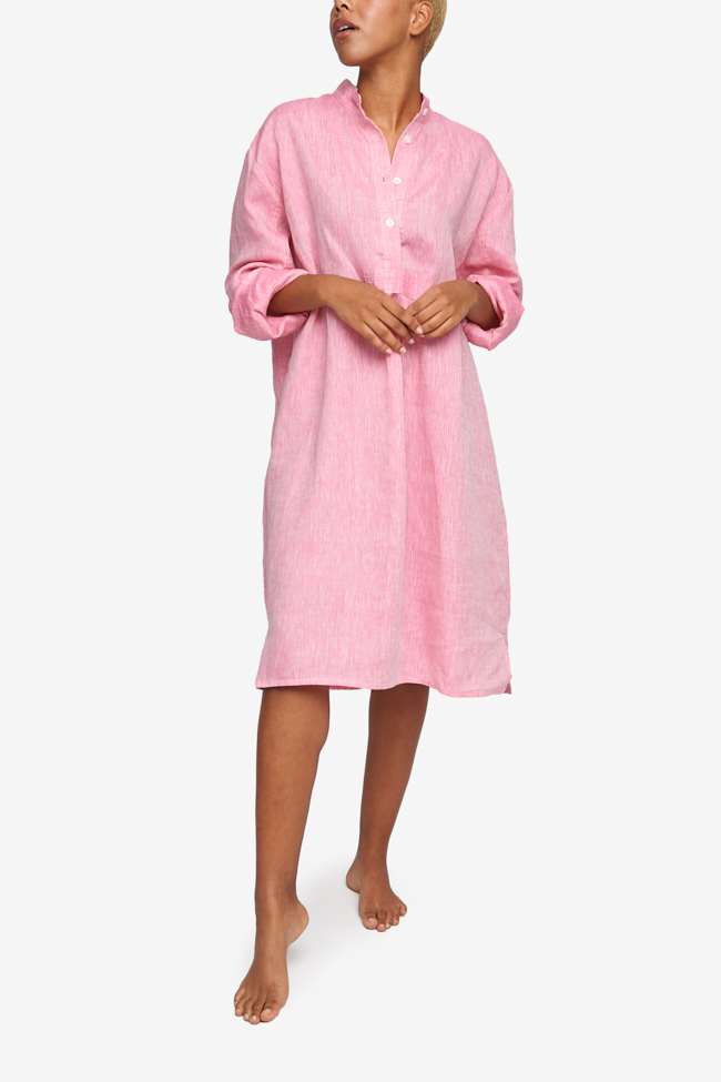 Full shot of a black woman with a blonde buzz cut, barefoot against a white background. She is wearing a below-the-knee-length nightshirt that has long, cuffed sleeves, a three-quarter placket with 4 buttons and a stand collar. It's made in a pink crisp, textured linen.
