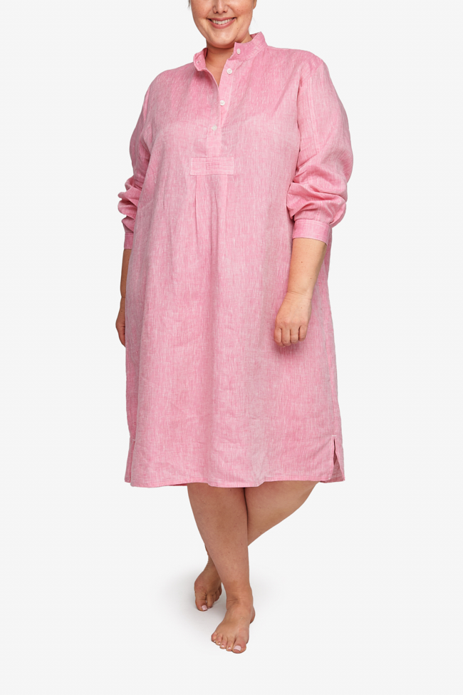 Full shot of a plus-sized white woman with her hair tied back in a low bun, barefoot against a white background. She is wearing a below-the-knee-length nightshirt that has long, cuffed sleeves, a three-quarter placket with 4 buttons and a stand collar. It's made in pink crisp, textured linen.