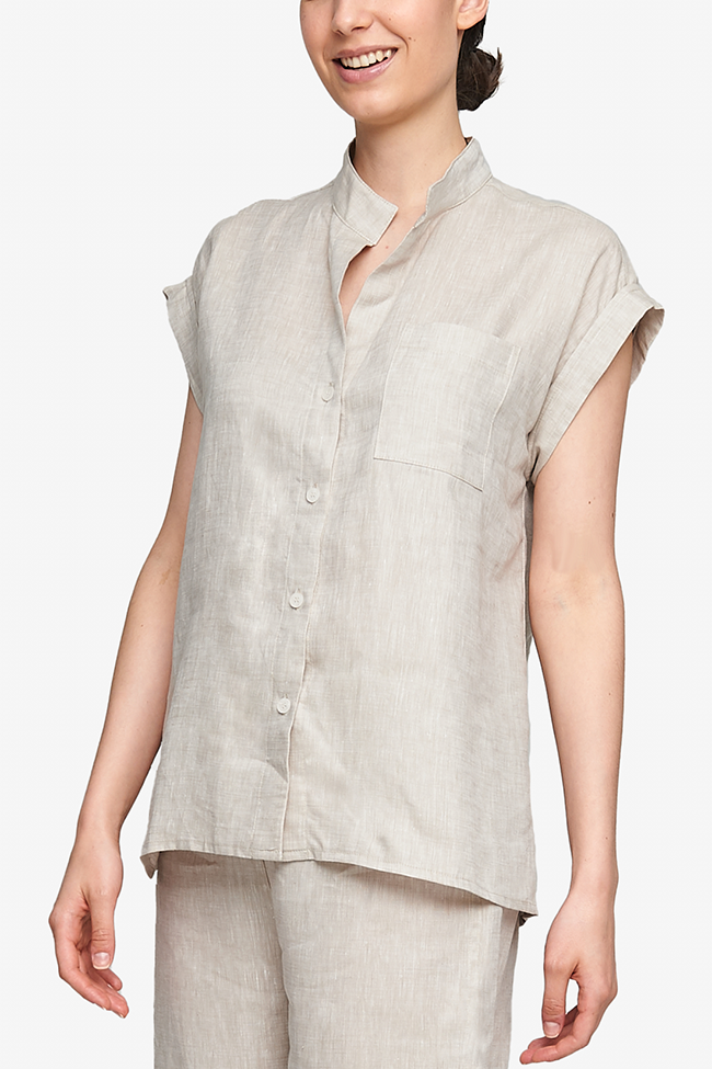 Cropped shot of a white woman, her torso is the focus. Wearing a pyjama top with a full placket and cuffed, cap sleeves. One breast pocket on the left side. It's made from a crisp linen in a cool, sandy beige colour.