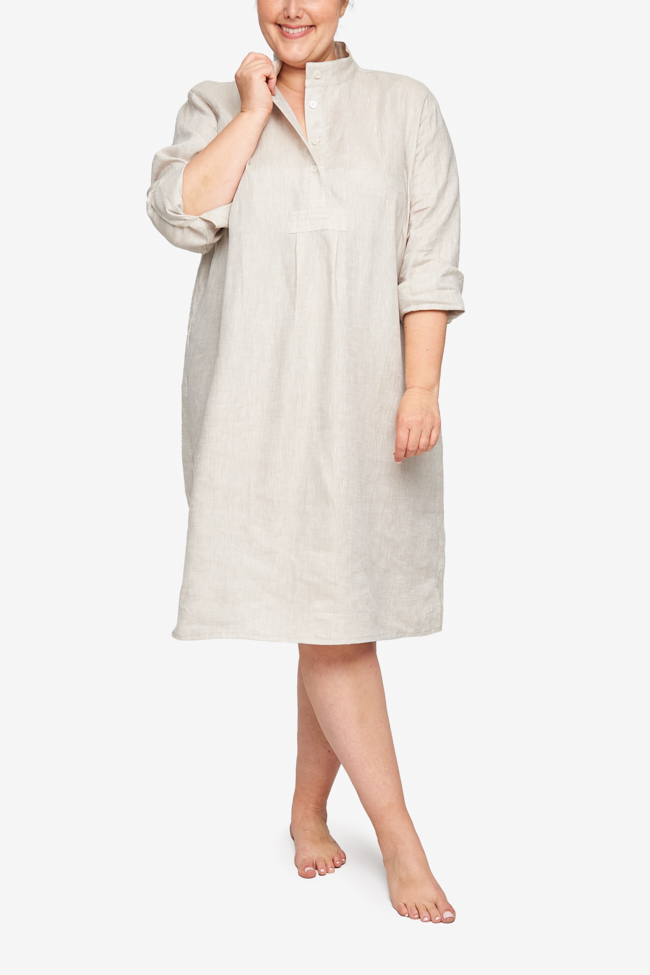 Full shot of a plus-size white woman with her hair tied back in a low bun, barefoot against a white background. She is wearing a below-the-knee-length nightshirt that has long, cuffed sleeves, a three-quarter placket with 4 buttons and a stand collar. It's made in cool beige, crisp, textured linen.