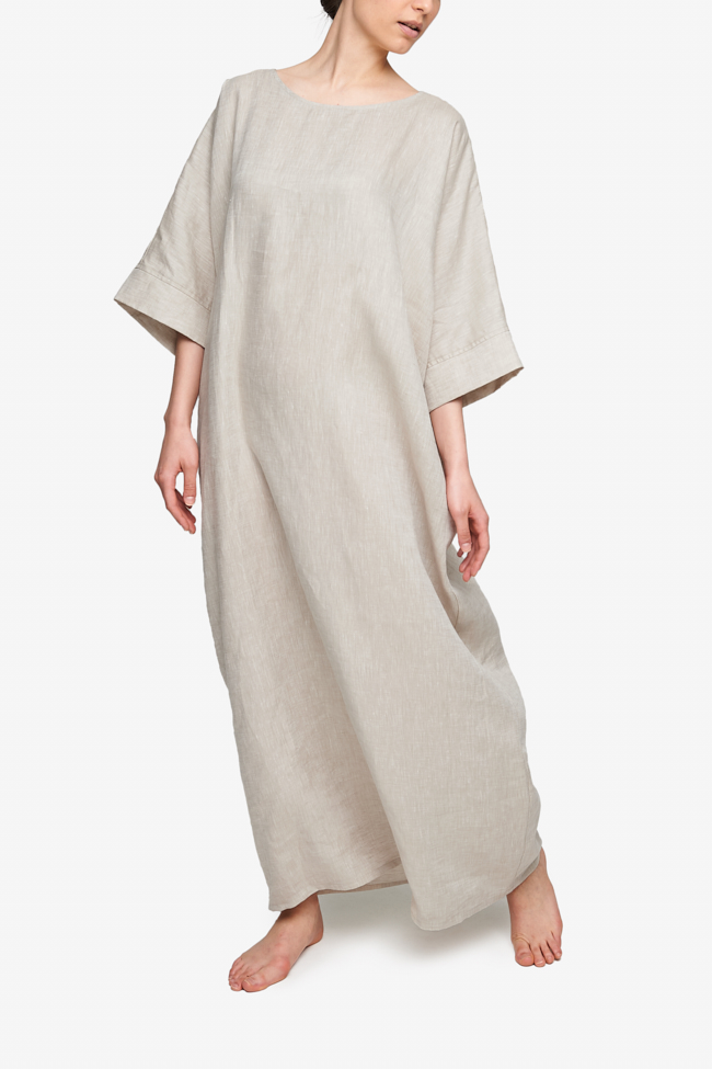 Full body shot of white woman, barefoot against a white background. She is wearing a floor length caftan that has wide, three quarter length sleeves, a boat neck and pockets. It has an overly generous amount of ease, especially in this hips giving it a draped, extra relaxed fit. Made in a cool sandy beige linen.