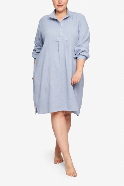 Full shot of a plus-size white woman with her hair tied back in a low bun, barefoot against a white background. She is wearing a below-the-knee-length nightshirt that has long, cuffed sleeves, a three-quarter placket with 4 buttons and a stand collar. It's made in silvery-blue cotton double gauze with a soft, loose weave.