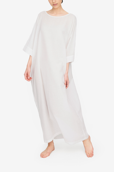 Full body shot of a white woman, barefoot against a white background. She is wearing a floor length caftan that has wide, three quarter length sleeves, a boat neck and pockets. It has an overly generous amount of ease, especially in this hips giving it a draped, extra relaxed fit. Our Milano Featherweight cotton linen blend is super lightweight and sheer. There is a subtle white on white horizontal stripe texture woven into the fabric.