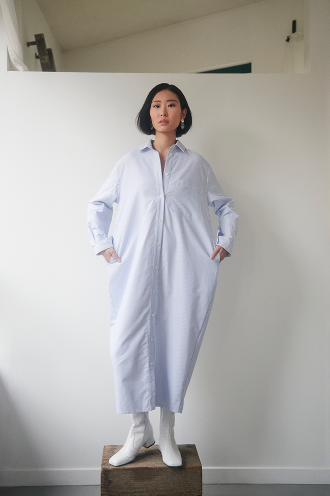 Xuan wears a long blue and white striped oxford cotton shirti with long sleeves and a button placket that opens all the way up.