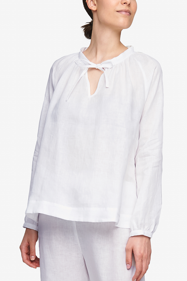 Cropped shot of a white woman, her torso is the focus. She is wearing a pyjama top with raglan sleeves with button cuffs, the hem hitting her at the hip. There is a little bow at the centre front of the gathered neck line. Made in our favourite, crisp white linen.