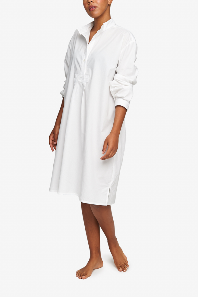 Full shot of a black woman with a blonde buzz cut, barefoot against a white background. She is wearing a below-the-knee-length nightshirt that has long, cuffed sleeves, a three-quarter placket with 4 buttons and a stand collar. It's made from a crips, lightweight cotton shirting.