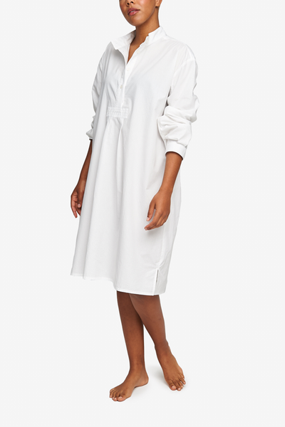 Full shot of a black woman with a blonde buzz cut, barefoot against a white background. She is wearing a below-the-knee-length nightshirt that has long, cuffed sleeves, a three-quarter placket with 4 buttons and a stand collar. It's made from a crips, lightweight cotton shirting.